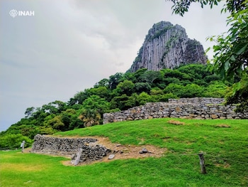 Montaña rocosa con formaciones columnares y vegetación, muros de piedra prehispánicos en laderas verdes, cielo nublado y logo de INAH