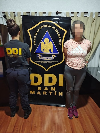 Oficial de policía de espalda y mujer con rostro pixelado de frente, ante un banner negro de la DDI San Martín de la Policía de Buenos Aires