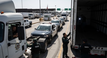 Agentes uniformados inspeccionan un camión de carga blanco en un cruce fronterizo. Hay patrullas, una garita, la bandera de México y el conductor del camión visible.