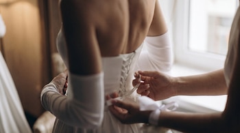 Vista trasera de una novia en un vestido blanco sin tirantes, mientras otra persona ata los cordones del corsé en su espalda. Las manos son visibles.