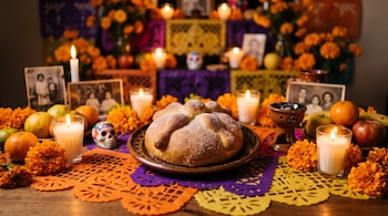 Pan espolvoreado con azúcar en plato de barro, rodeado de flores de cempasúchil, velas encendidas, frutas y papel picado.
