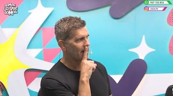 Christian Meier viste camiseta negra y coloca un dedo sobre sus labios frente a un fondo colorido con logotipos de Zaca TV