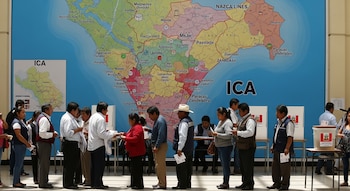 Varias personas en una fila dentro de un lugar de votación, con un mapa grande y detallado de la región de Ica, Perú, y uno pequeño de Ica al fondo.