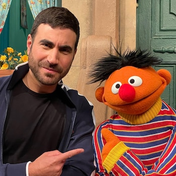 Un hombre sonríe y señala a un títere naranja con pelo negro, ojos grandes y nariz roja, posando frente a una puerta verde y flores amarillas