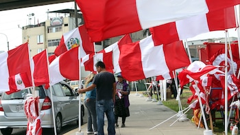 Múltiples banderas de Perú de color rojo y blanco, algunas con el escudo nacional, ondean sobre un grupo de personas en una vereda junto a un auto plateado