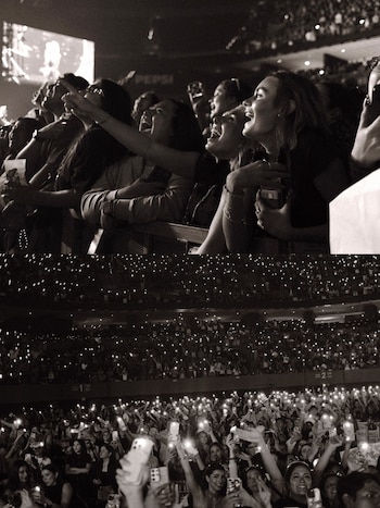 Dos imágenes en blanco y negro de asistentes sonriendo y levantando teléfonos con luces encendidas en un concierto masivo