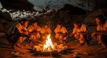 Seis figuras de Homo erectus sentadas en cuclillas alrededor de una hoguera brillante dentro de una cueva oscura, con rocas y tierra visibles.