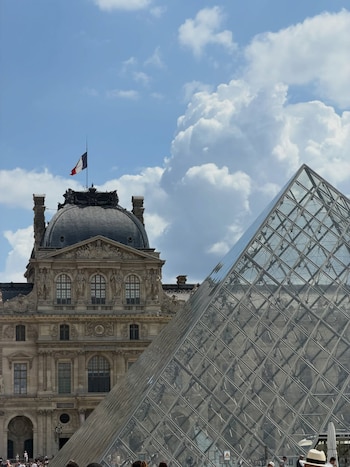 Pirámide de cristal del Louvre en primer plano junto al edificio histórico del Museo del Louvre, bandera francesa, cielo azul con nubes