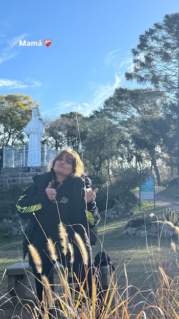 Mujer con chaqueta negra, pulgares arriba, mueca en el rostro. Estatua blanca, árboles, cielo azul, nubes, pastos. Texto "Mamá ❤️"