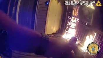 Imagen de cámara corporal mostrando el brazo extendido de un agente de policía mientras un violento incendio consume una estructura residencial a la derecha