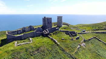 Ruinas de piedra, incluyendo dos torres, sobre una colina verde con muros y un camino. Un vehículo azul y dos personas cerca del mar y el cielo claro.