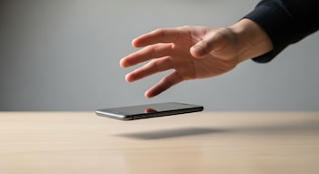 Una mano de piel clara está a punto de soltar un teléfono inteligente negro sobre una mesa de madera clara, con un fondo gris neutro.