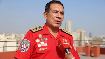 Un hombre con uniforme rojo de bombero, insignias doradas, un parche "BOMBEROS PERU" y "C. PAREDES A.", y un logo en la manga, habla a un micrófono rojo de Andina