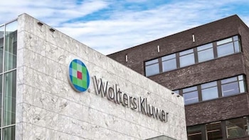 Fachada de un edificio de mármol claro con el logo y el nombre de Wolters Kluwer en relieve, con un edificio de ladrillo oscuro y ventanas detrás y un cielo nublado