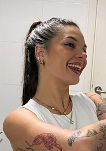 Primer plano de una mujer joven con cabello oscuro recogido, sonriendo hacia la derecha, con aretes y collar dorados. Se aprecian tatuajes en sus brazos