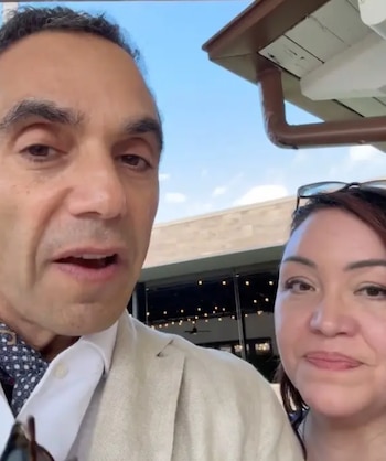 Primer plano de un hombre hablando junto a una mujer, ambos al aire libre con un cielo azul y parte de una estructura de edificio visible