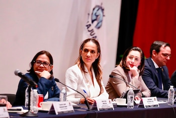 Cuatro personas sentadas en una mesa de presídium con micrófonos, botellas de agua e identificadores de nombre ante un telón blanco y rojo