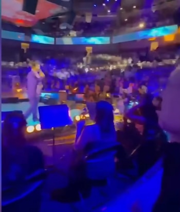 Un cantante de pie en un escenario bajo luces azules, frente a una gran audiencia en un recinto. Se ven siluetas de personas en primer plano