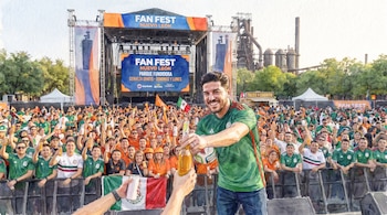 Un hombre sonriente en camiseta de México entrega una botella. Gran multitud de asistentes, banderas mexicanas, un escenario y estructuras industriales. Estilo acuarela.