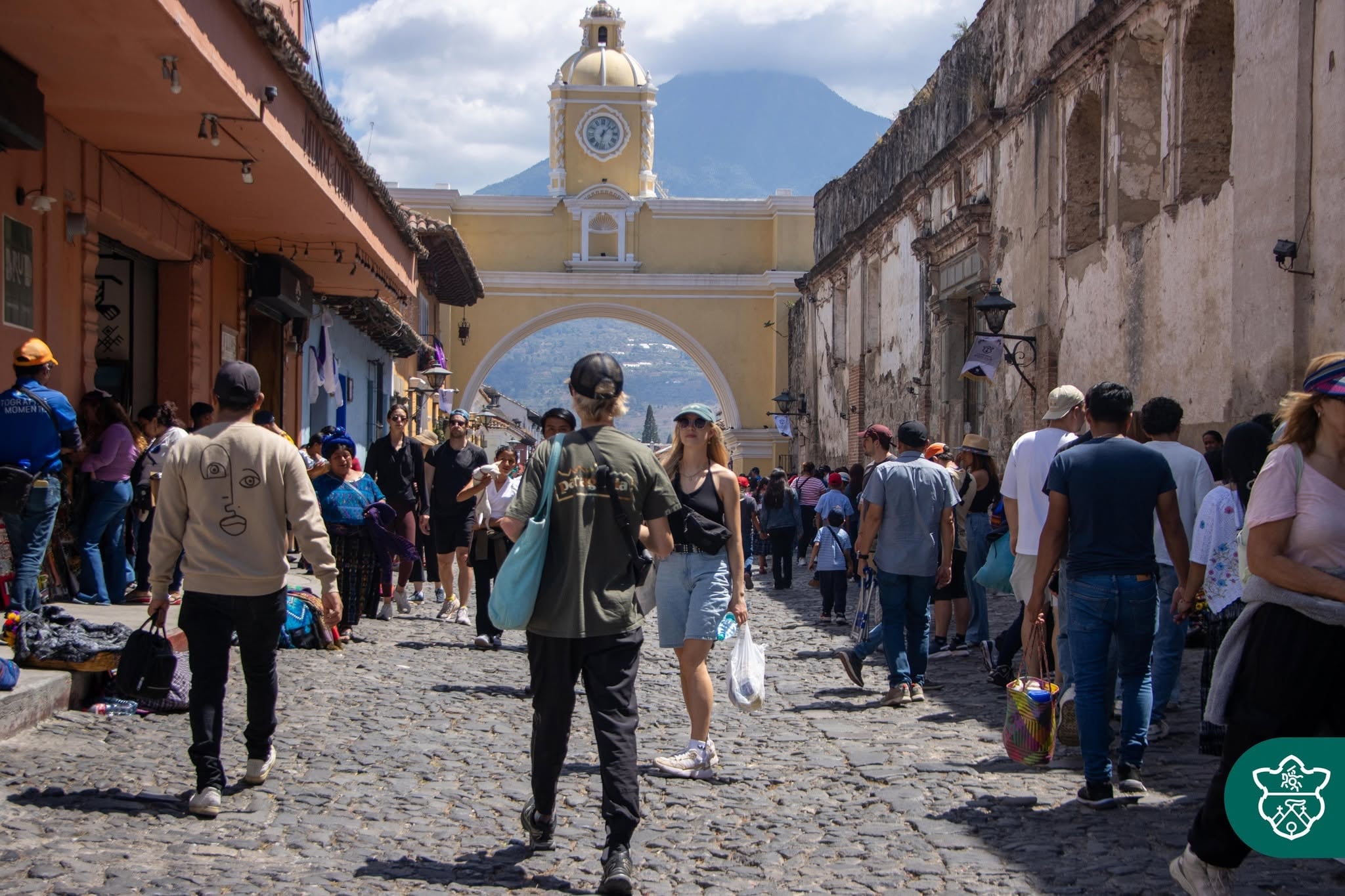 Una concurrida calle empedrada en Antigua Guatemala muestra a turistas y locales paseando bajo el icónico Arco de Santa Catalina, con un volcán majestuoso en el horizonte. (Municipalidad de Antigua Guatemala)