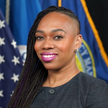 Primer plano de La’ Toya Prieur, una mujer sonriente con trenzas largas y un lado rapado, vestida con un traje oscuro, frente a banderas