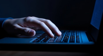 Plano cerrado de una mano pálida apoyada en el teclado de una laptop, iluminada por una luz azul fría que emana de la pantalla en un entorno oscuro.