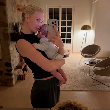 Mujer rubia con un moño alto sosteniendo a un bebé mientras se toma una selfie en un espejo. El fondo muestra una sala de estar con ventana y muebles modernos