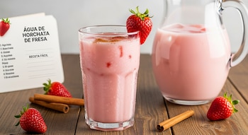 Primer plano de un vaso de horchata de fresa con una fresa en el borde, una jarra, más fresas, palitos de canela y una tarjeta de receta sobre madera.