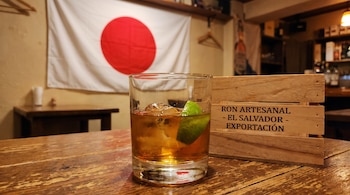 Un vaso de ron con hielo y lima en una mesa de madera. Al fondo, una bandera de Japón y una caja con el texto "RON ARTESANAL - EL SALVADOR - EXPORTACIÓN".