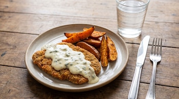 Un plato de milanesa empanada con salsa cremosa de Roquefort y batatas asadas, junto a un vaso de agua, un cuchillo y un tenedor sobre una mesa de madera.