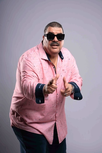 Hombre con gafas de sol oscuras y bigote, con camisa rosa de manga larga estampada, que apunta al frente con ambos índices