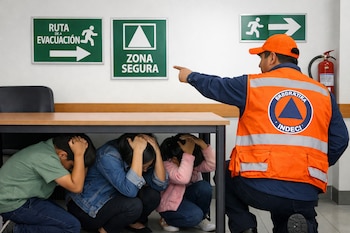 En un simulacro sísmico, personas se cubren bajo una mesa mientras un brigadista de INDECI con chaleco naranja señala una ruta de evacuación.