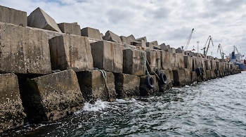 Bloques de hormigón apilados junto al agua con neumáticos colgados, grúas portuarias al fondo y un barco en la superficie marina.