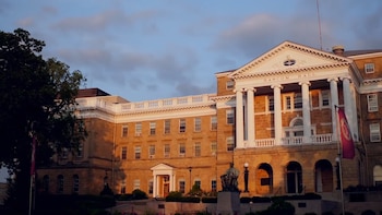 Fachada de un edificio de piedra con columnas blancas, la inscripción Bascom Hall, una estatua en la entrada y banderas laterales