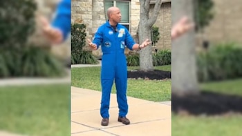 Un astronauta calvo, vistiendo un traje de vuelo azul con parches, se encuentra al aire libre con los brazos abiertos, frente a una casa de piedra y vegetación