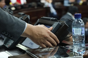 Mano de persona con traje presiona lector biométrico negro sobre un atril de madera. Un micrófono y una botella de agua están sobre la mesa