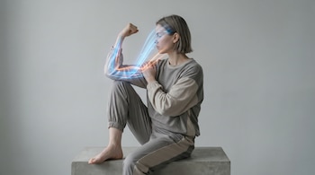 Figura de género ambiguo sentada en ropa deportiva gris, flexionando un brazo. Rayos de luz azul y naranja fluyen desde su bíceps hacia la cabeza.