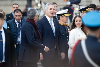 Hombre de traje azul y corbata clara, oficial militar uniformado y mujer de abrigo blanco sonrientes, en un evento público formal