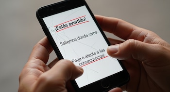 Primer plano de dos manos sosteniendo un celular negro con pantalla rota. En la pantalla se leen mensajes de extorsión en español.