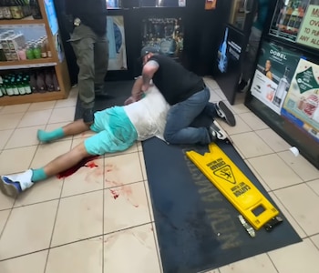 Hombre con camiseta blanca y pantalón corto turquesa yace en el suelo de baldosas, con sangre en las piernas, mientras otro hombre lo sujeta y un tercero está de pie