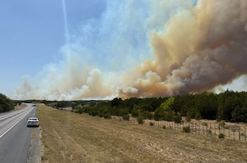 Una autopista con un vehículo blanco en el arcén junto a un área arbolada y densas columnas de humo que se elevan al cielo
