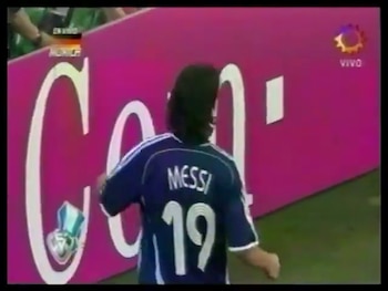 Vista trasera de Lionel Messi vistiendo la camiseta número 19 de Argentina con cabello largo, parado en un campo de fútbol frente a una valla publicitaria rosa
