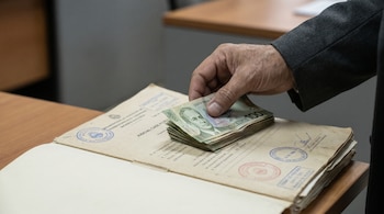 Mano adulta colocando billetes de cien colones costarricenses sobre un expediente judicial abierto con sellos visibles sobre una mesa de madera.