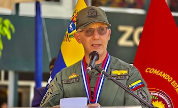 Un militar con uniforme verde de Venezuela, gorra, anteojos y condecoración habla ante un micrófono frente a dos banderas