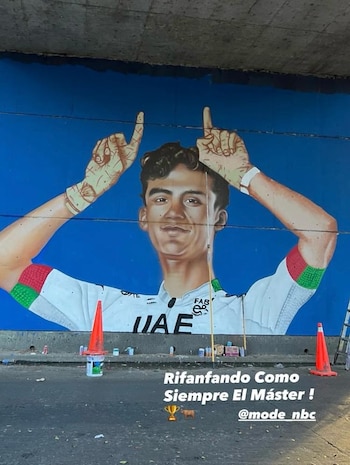 Un mural de un ciclista con camiseta blanca del equipo UAE Team Emirates y brazos levantados. En el suelo, conos de tráfico y latas de pintura