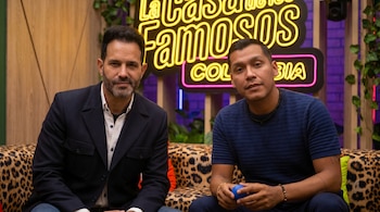 Dos hombres, Alejandro Estrada y Juanda Caribe, sentados en un sofá de estampado de leopardo, con un logo de neón "La Casa de los Famosos Colombia" de fondo.