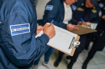 Un agente con una insignia que dice El Salvador en la manga escribe en una hoja sobre un portapapeles de madera junto a otros colegas