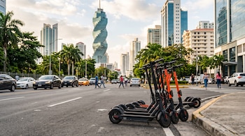 Varios scooters eléctricos negros y naranjas en la orilla de una calle, con automóviles, peatones y rascacielos al fondo.