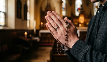 Primer plano de manos de un hombre joven unidas en oración, sosteniendo un rosario de madera con un crucifijo, dentro de una iglesia
