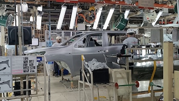 Trabajadores en cascos blancos ensamblan una pickup plateada en la línea de producción de una fábrica de automóviles con luces y maquinaria industrial